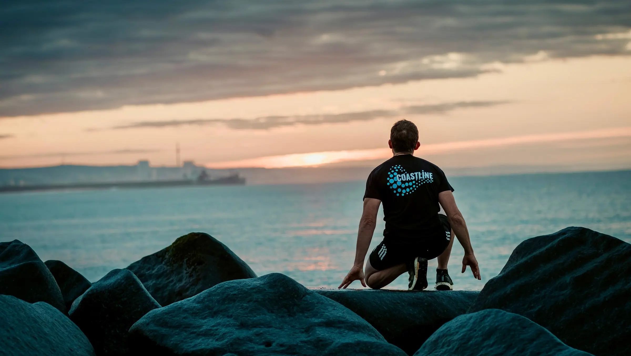 Mark crouches on the sea defence rocks with his back to the camera, looking out to sea at sunset.