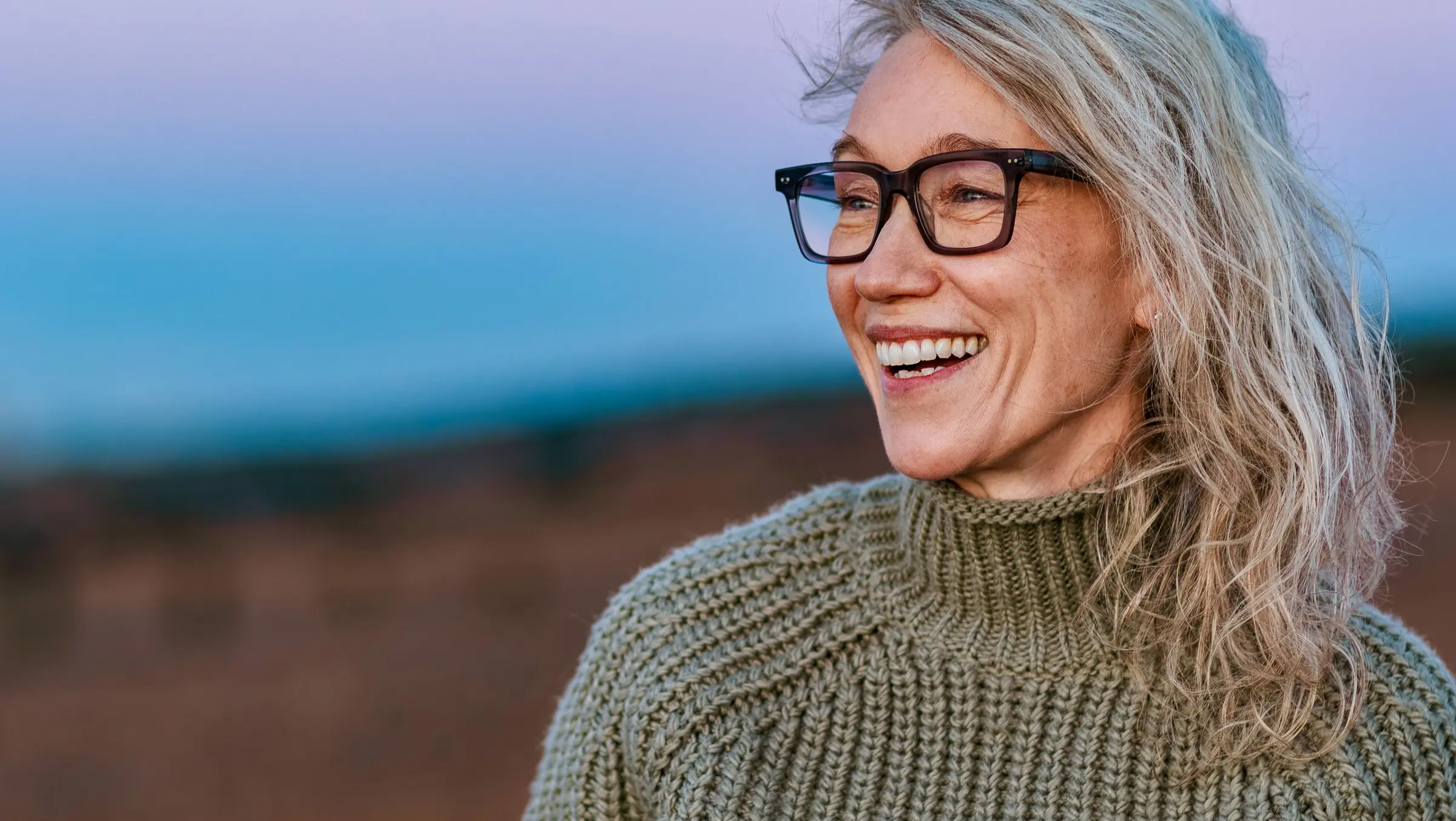 Julie, in glasses and a green knitted jumper, laughs and looks along the beach at dusk.